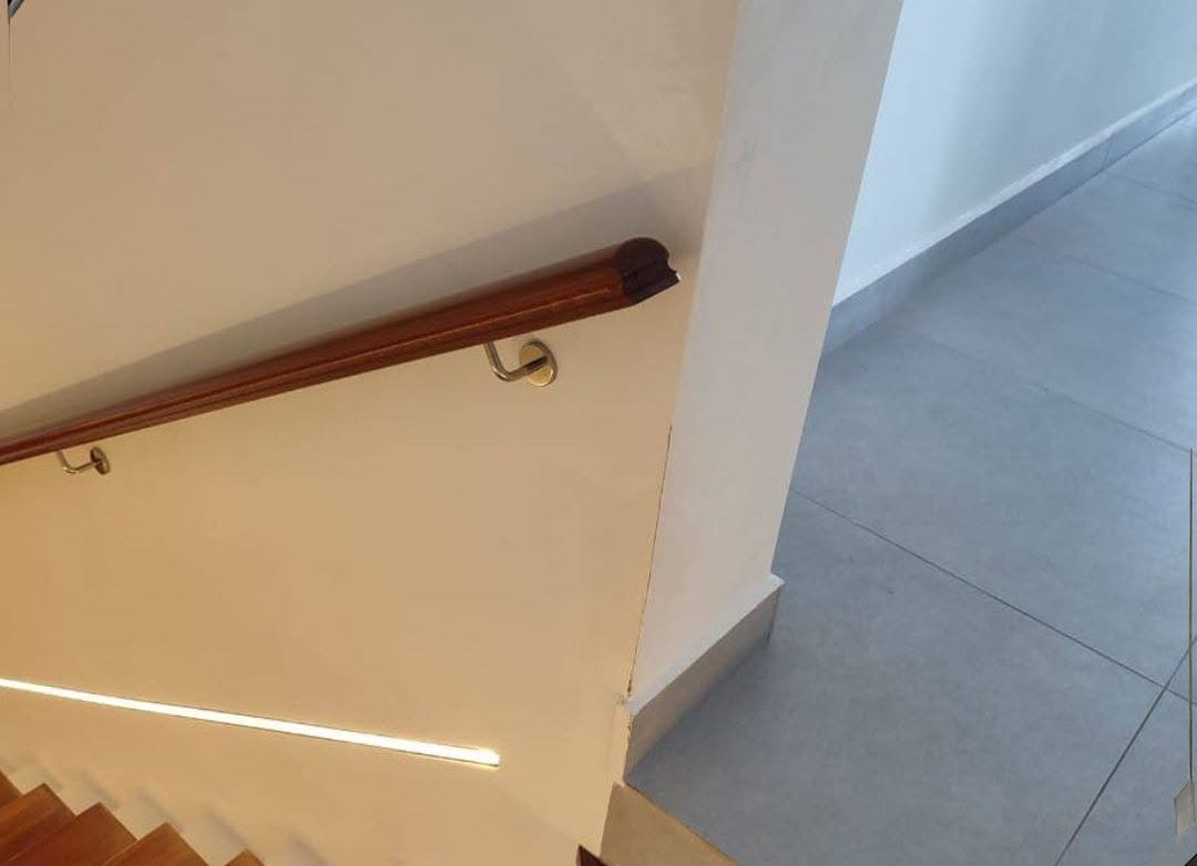 A dark timber wall handrail following a staircase, with a lit line recessed in the stringer wall below it.