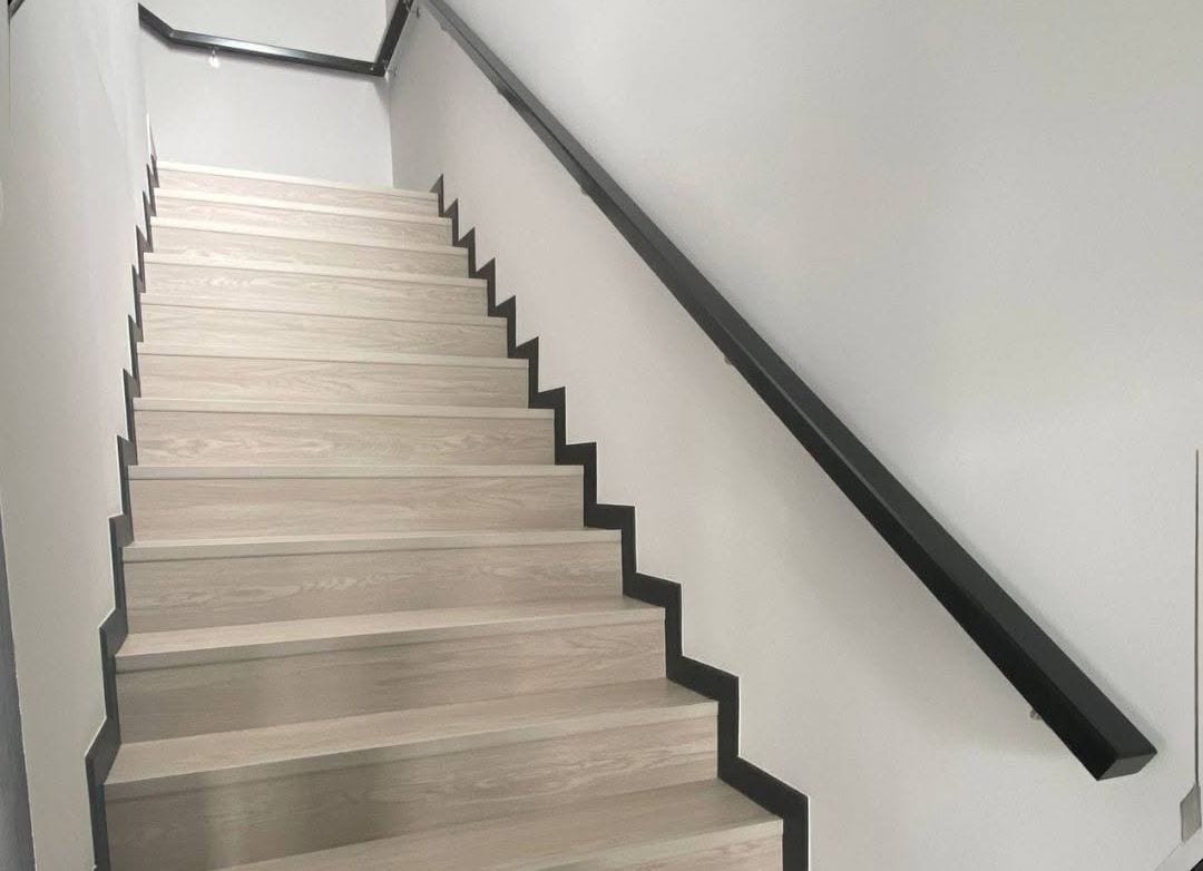 A slim flat-section black steel wall handrail running the full length of a staircase, against white plaster and pale timber treads.